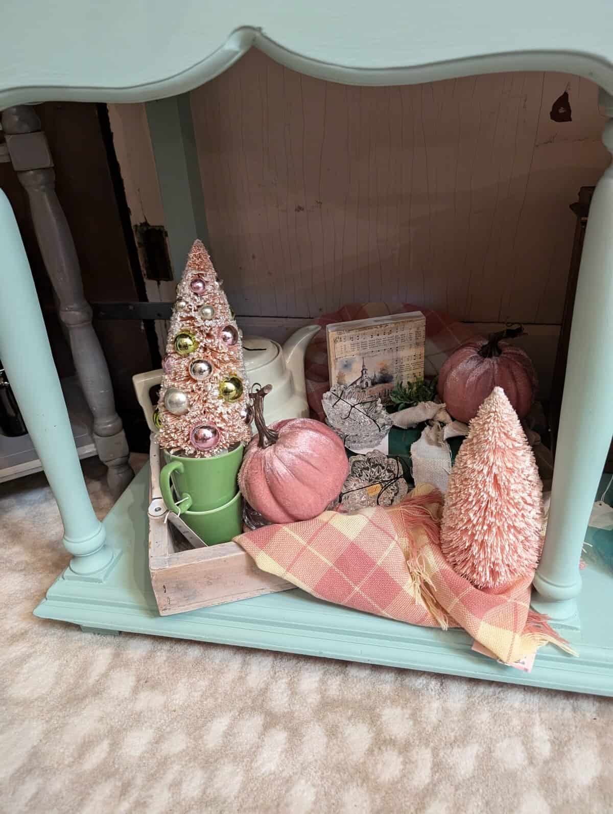A decorated shelf with pink bottle brush trees, pumpkins, a green mug, a plaid cloth, and various decorative items arranged on a blue wooden base.
