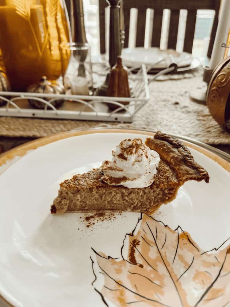 A slice of pie topped with whipped cream and a sprinkle of cinnamon sits on a plate with a leaf design; table decor is visible in the background.