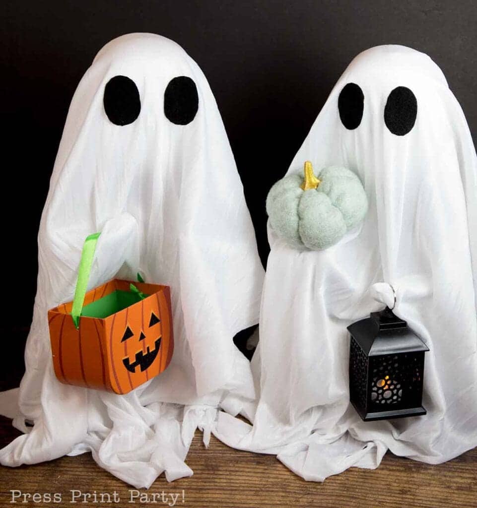 Two people are dressed as ghosts with white sheets and black eye cutouts; one holds a pumpkin-shaped basket, and the other holds a lantern and a small pumpkin.