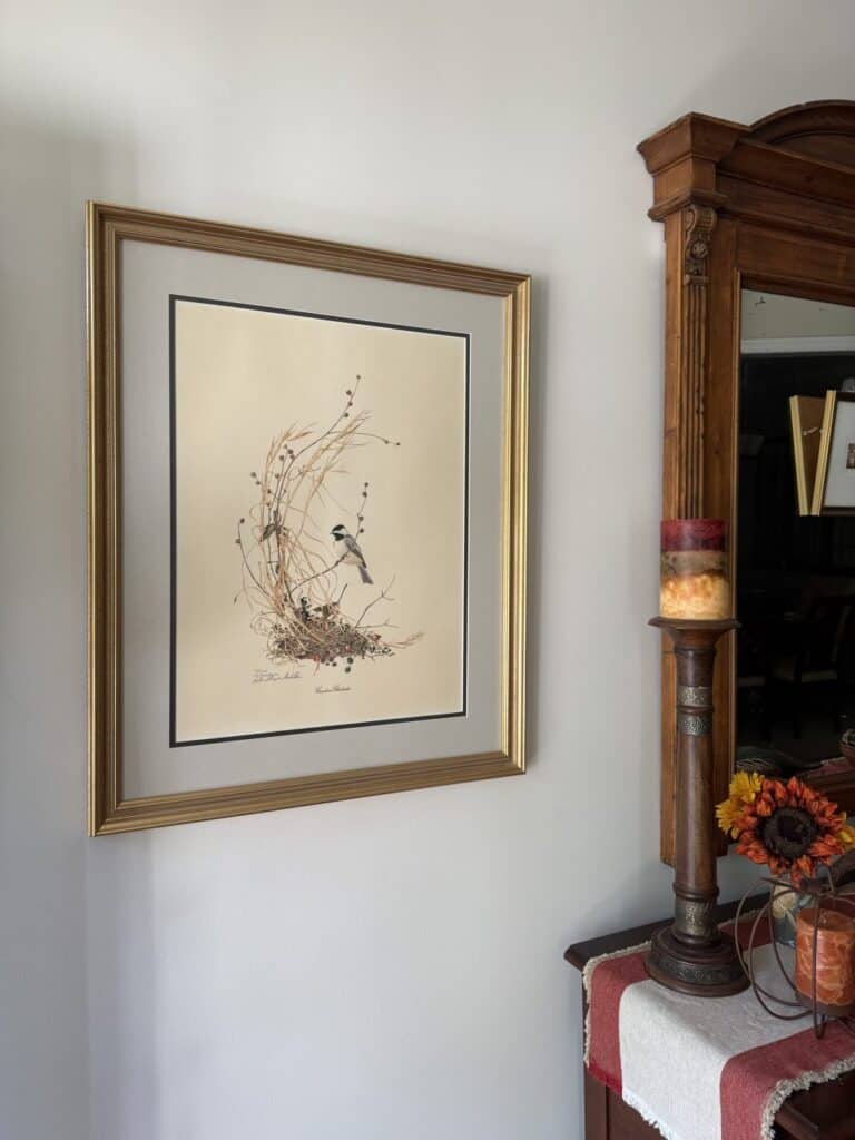 Framed botanical illustration with a bird hangs on a white wall next to a wooden mirror above a table with a candle and artificial flowers.