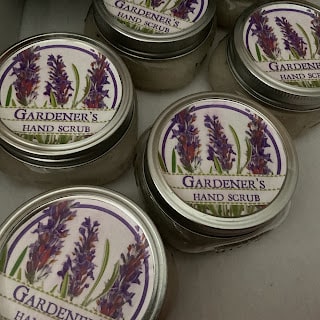 Five small jars labeled "Gardener's Hand Scrub" with purple flower illustrations on the lids are arranged closely together.