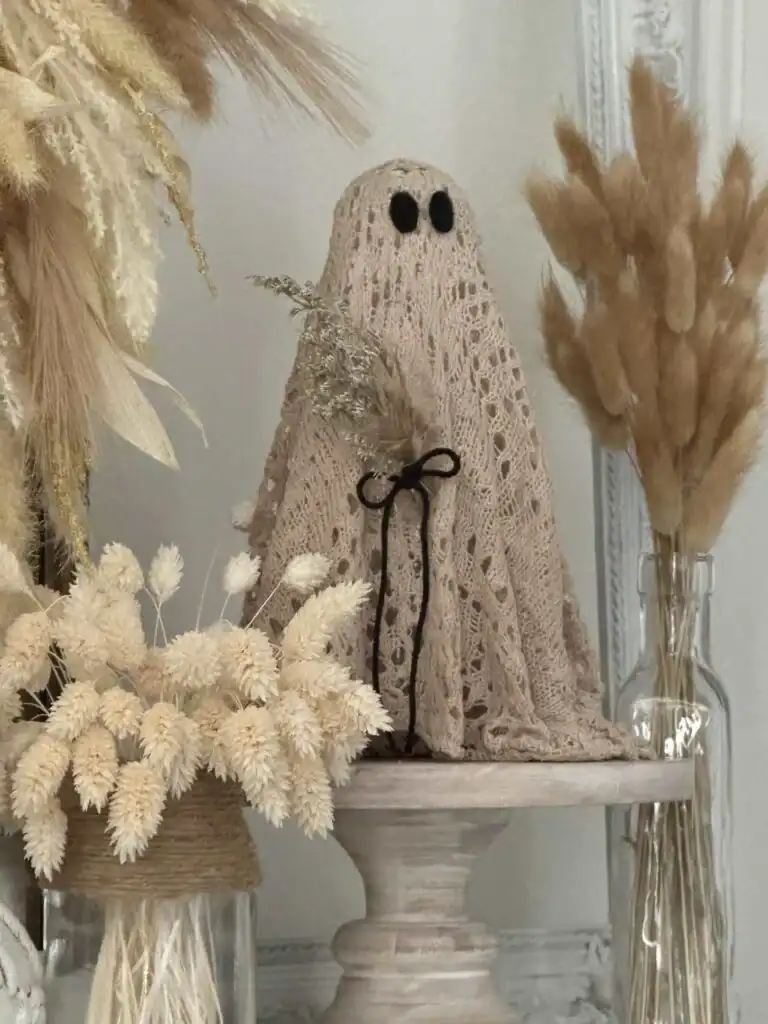 A small ghost figure with black eyes made from a lacy beige cloth stands on a pedestal, surrounded by dried beige and tan grasses in vases.