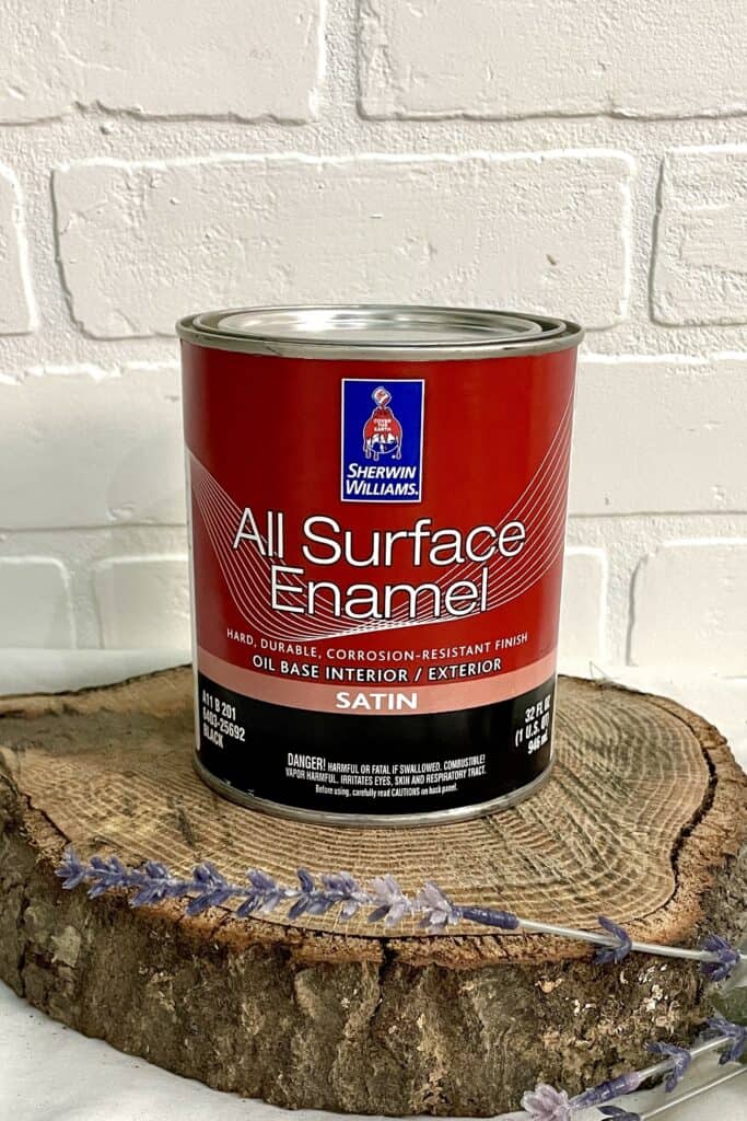 A can of Sherwin-Williams All Surface Enamel sits on a wooden slab with a white brick background and some lavender in the foreground.