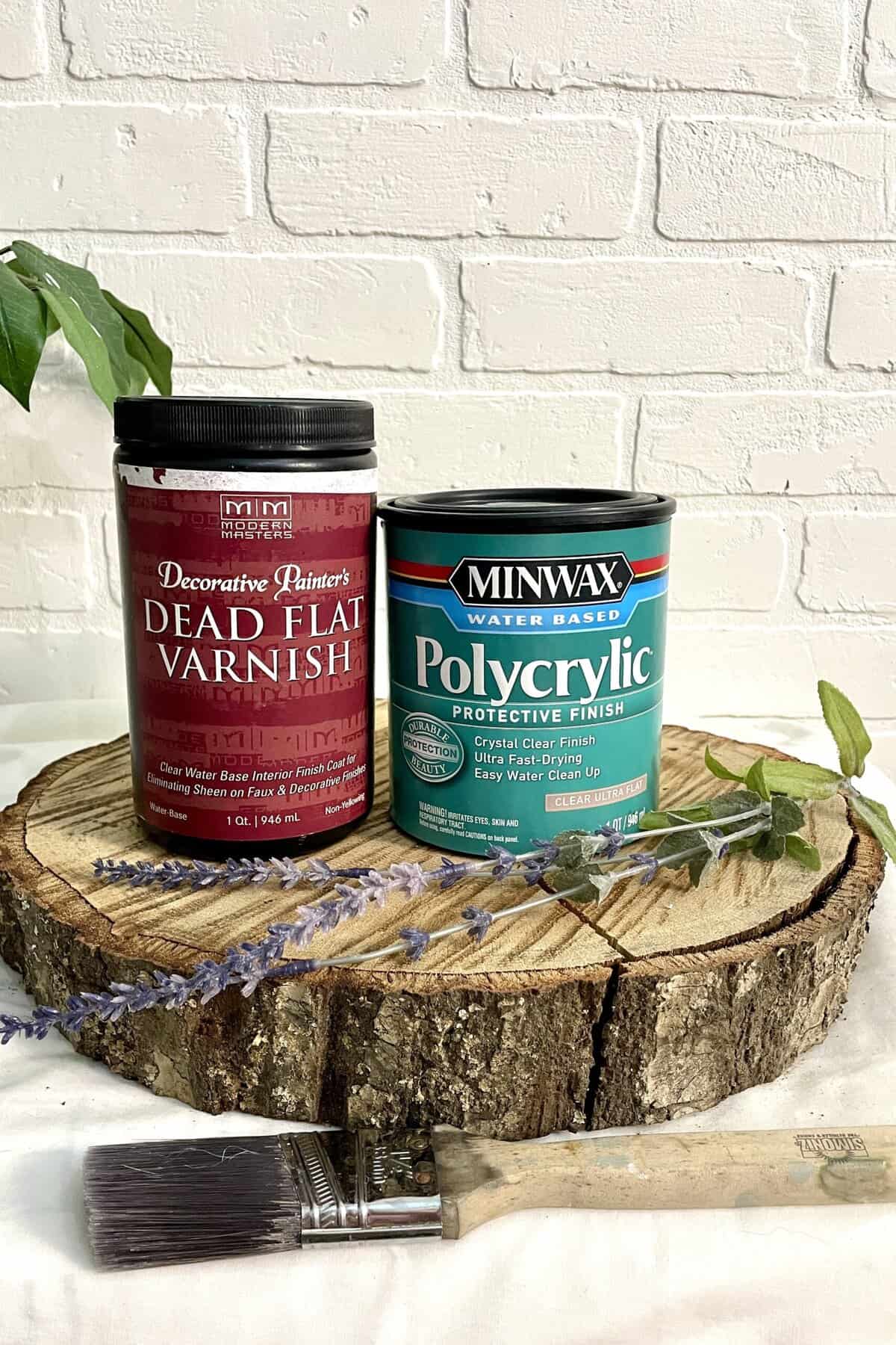 Two cans of wood finish—one Dead Flat Varnish and one Polycrylic—sit on a wooden slab, with a paintbrush and sprigs of lavender in front, against a white brick background.