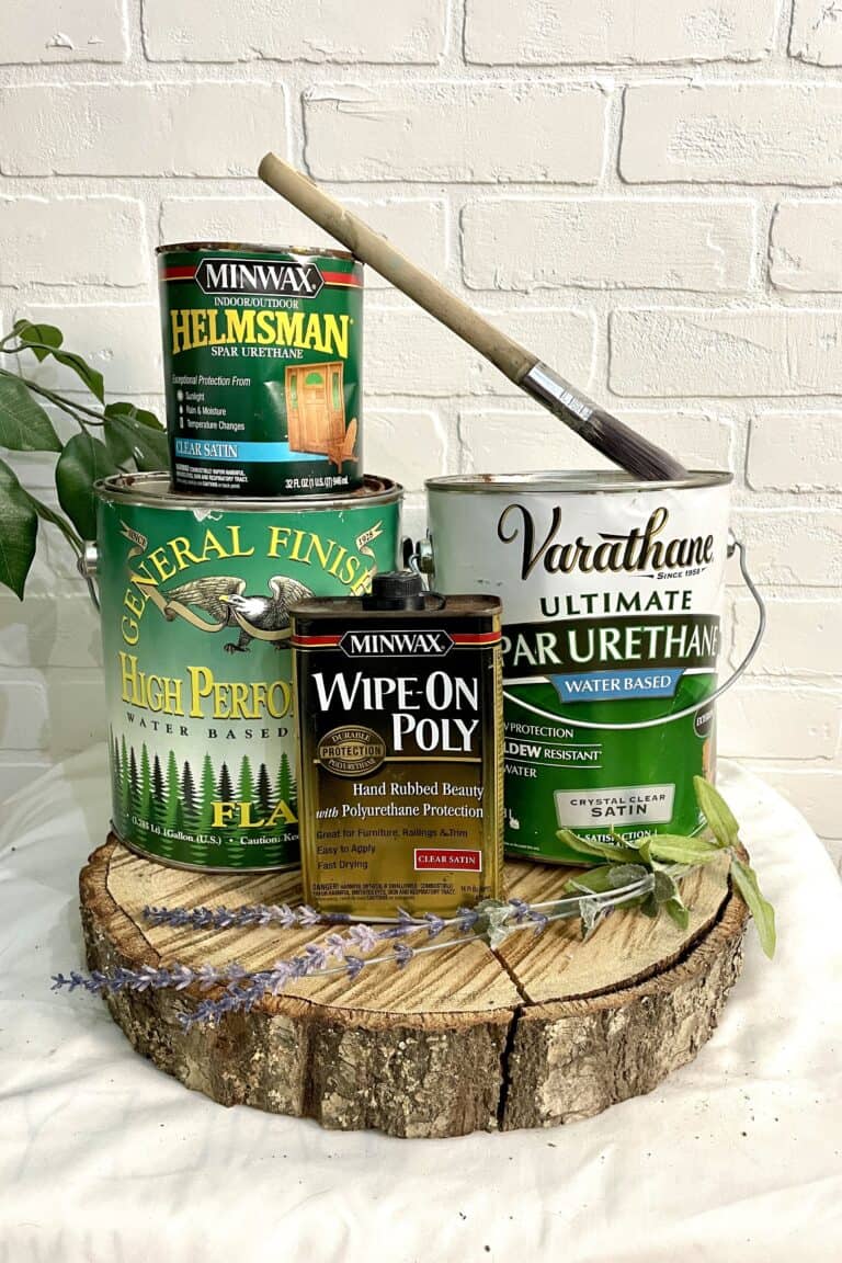Four cans of wood finish and a brush are arranged on a wooden slab against a white brick wall, with small greenery and lavender sprigs placed around them.