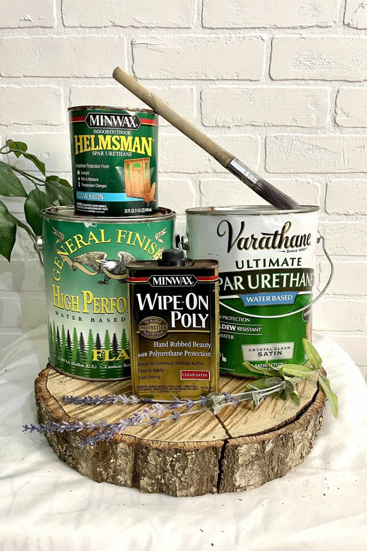 Four cans of wood finish and a brush are arranged on a wooden slab against a white brick wall, with small greenery and lavender sprigs placed around them.