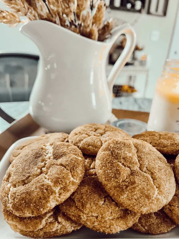 A plate of round, golden-brown cookies sits in front of a white ceramic pitcher with dried wheat and a lit candle on a table.