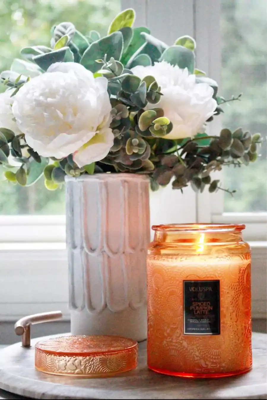 A lit orange candle in a textured glass jar sits on a marble surface next to its lid, with a white vase of artificial white flowers and greenery in the background.
