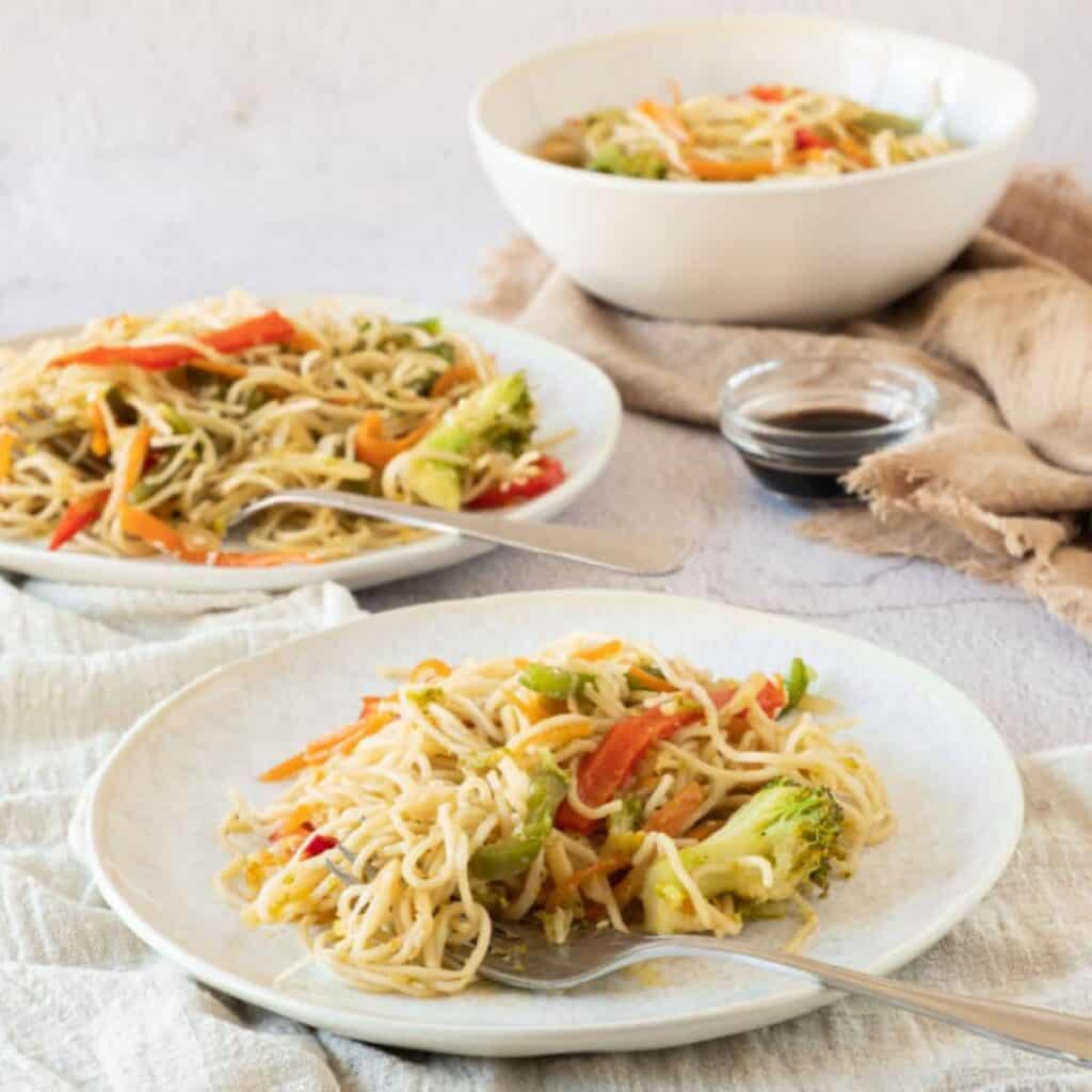 Two plates and a bowl filled with stir-fried noodles, mixed vegetables, and a small dish of sauce on a light-colored table.