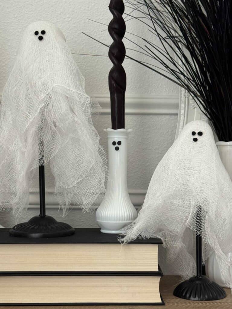 Two small ghost decorations made of white fabric sit on stands next to books and a vase, with a dark twisted candle and decorative black branches in the background.