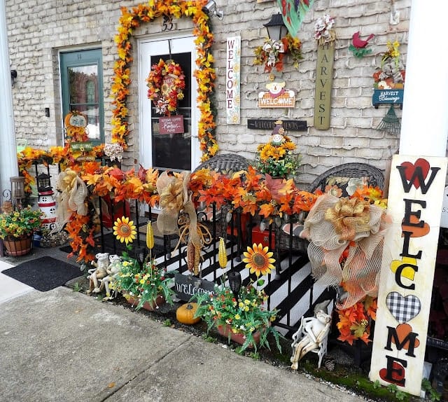 Front porch decorated for autumn with orange and yellow leaves, sunflowers, wreaths, welcome signs, scarecrows, and fall-themed ornaments.
