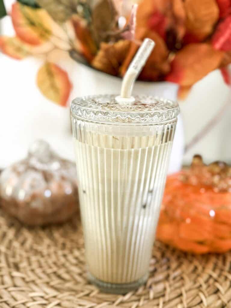 A glass of iced coffee with a lid and straw sits on a woven surface, with decorative pumpkins and autumn leaves in the background.