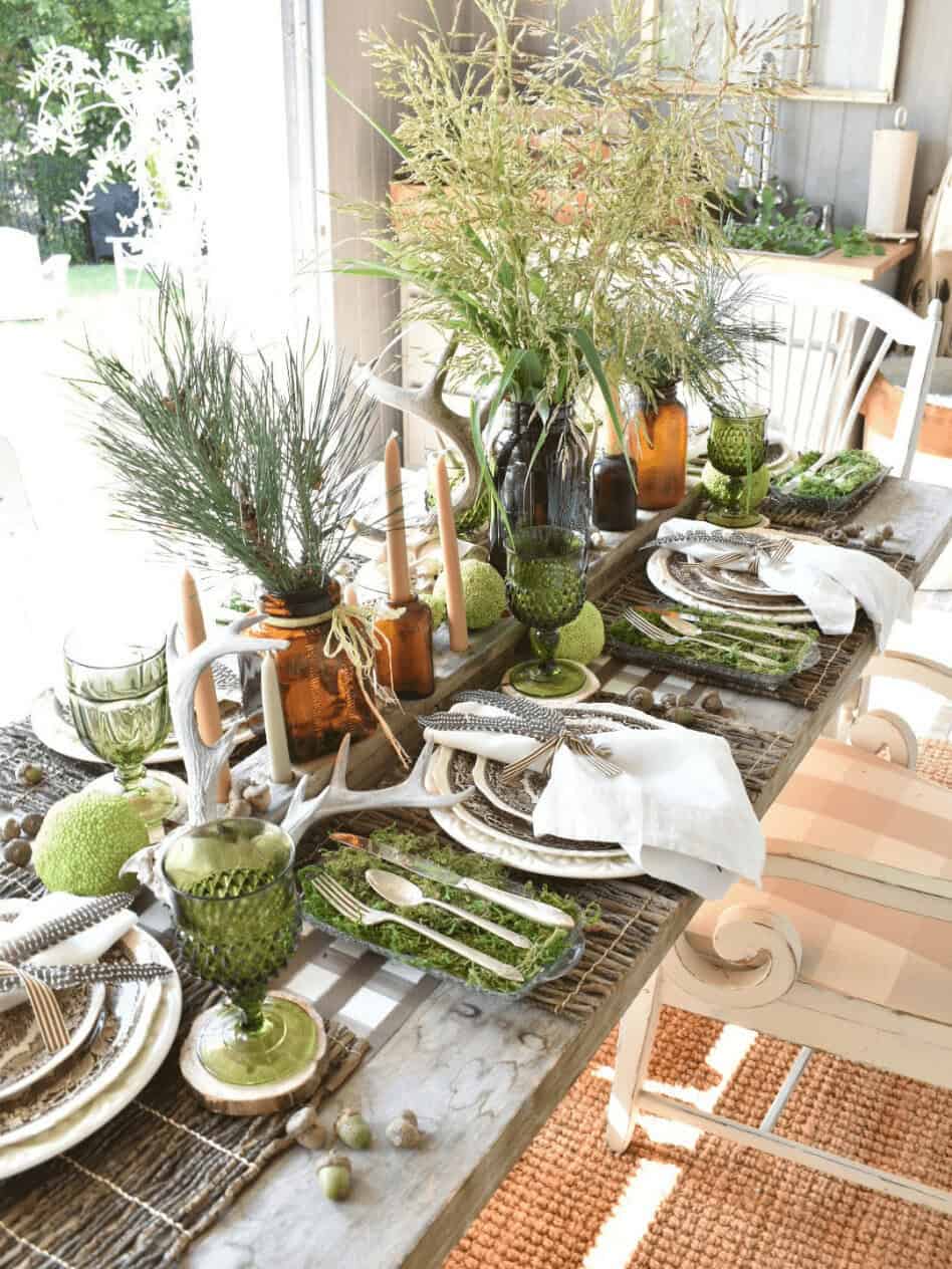 A rustic dining table set with green glassware, plates, brown bottles as vases, white napkins, candles, and natural greenery as centerpieces.