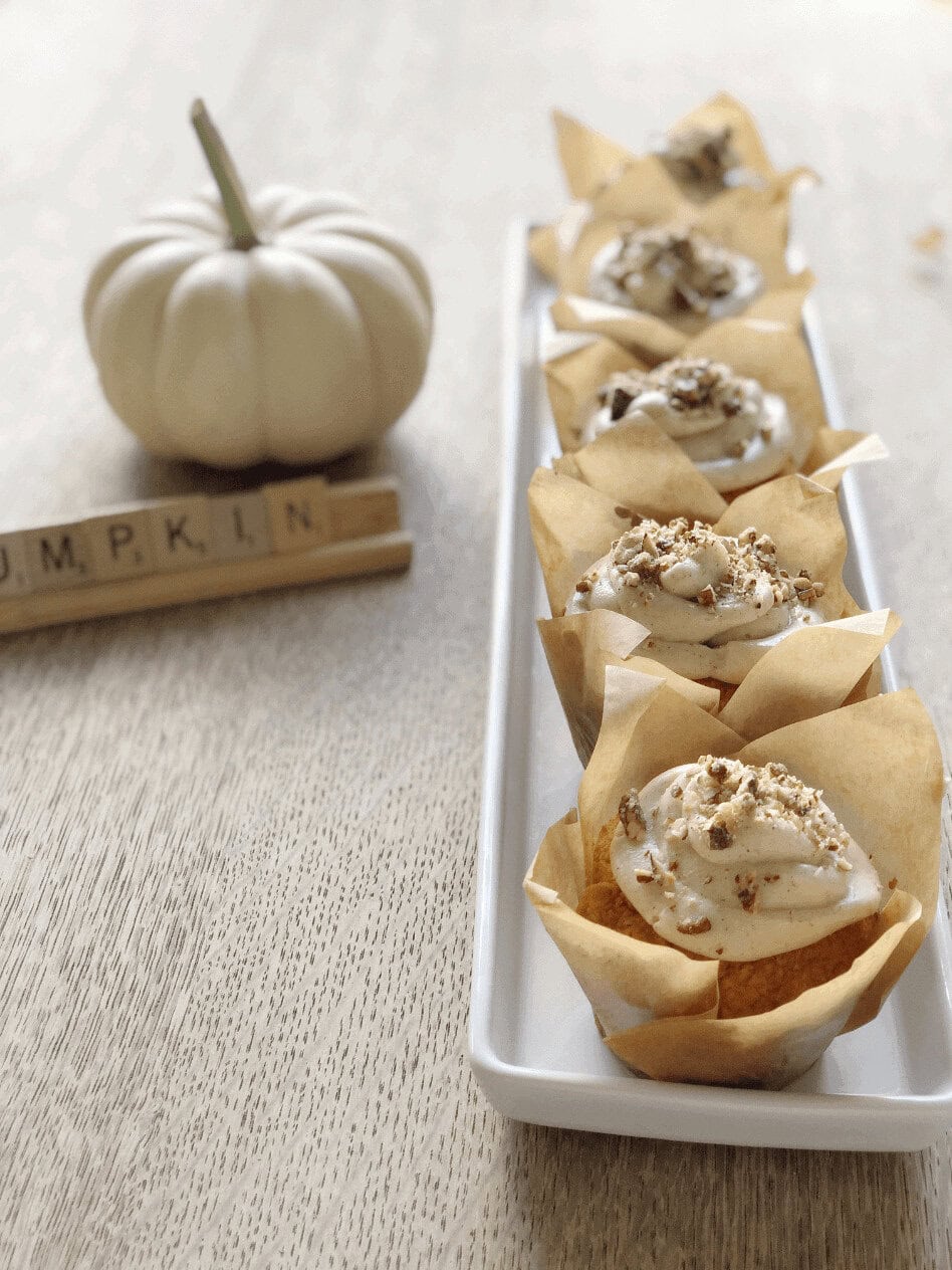 A row of cupcakes with frosting and nuts on a rectangular plate, next to a white pumpkin and Scrabble tiles spelling “PUMPKIN” on a wooden table.