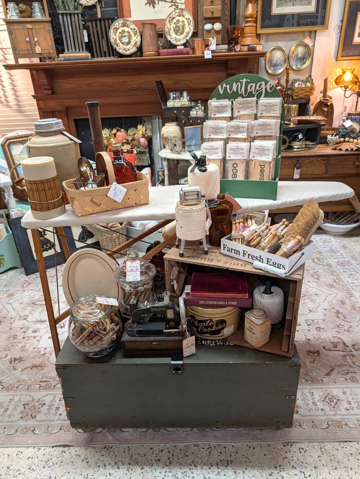 A display of vintage items including kitchenware, decor, and collectibles arranged on and around a wooden trunk and an ironing board in an antique shop.