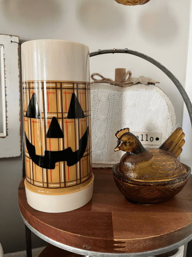 A ceramic mug with a plaid pattern and jack-o’-lantern face sits next to a small brown glass hen dish on a wooden shelf.