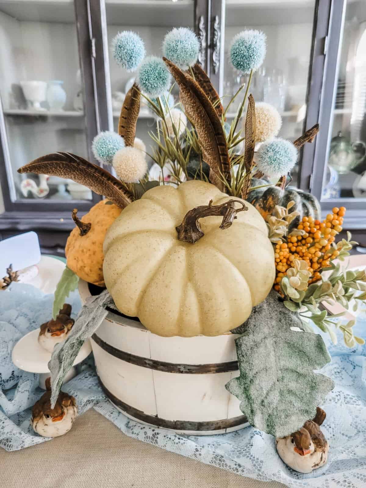 A white wooden bucket holds a light green pumpkin, yellow berries, green leaves, brown feathers, and blue and white pom-pom flowers as a decorative centerpiece.