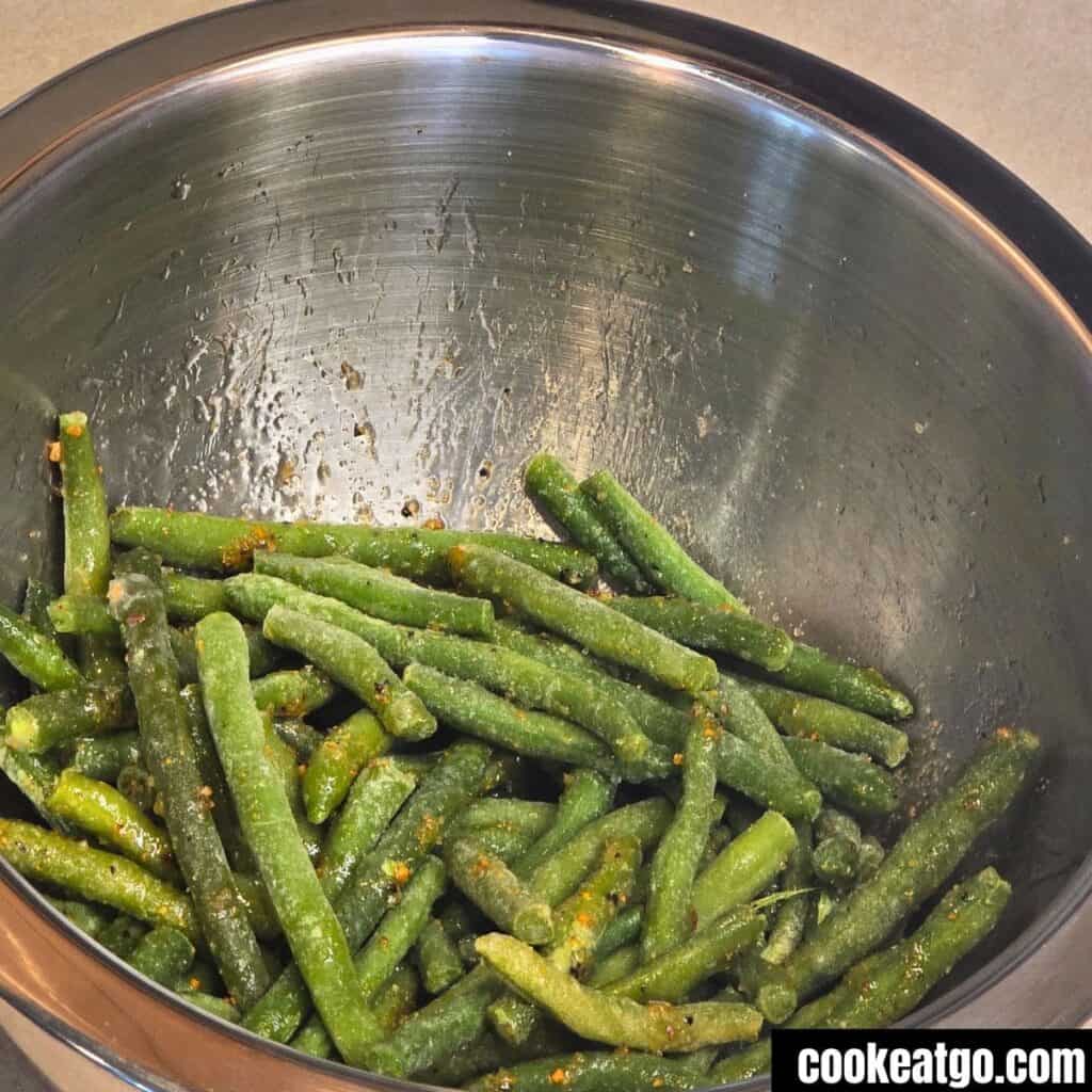 A stainless steel bowl containing seasoned frozen green beans with visible spices, set on a countertop. The image has a watermark: cookeatgo.com.