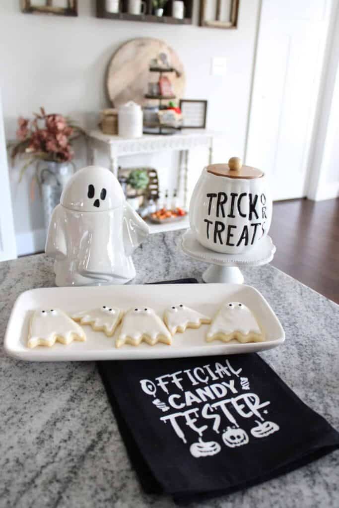 A kitchen counter displays ghost-shaped cookies on a plate, Halloween-themed containers, and a black towel labeled "Official Candy Tester.