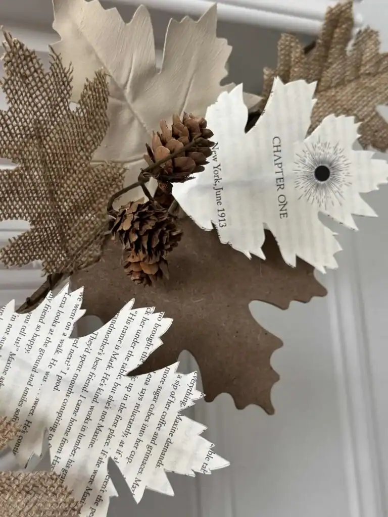 A decorative arrangement of artificial leaves made from book pages, burlap, and brown paper, accented with pinecones, displayed against a white background.
