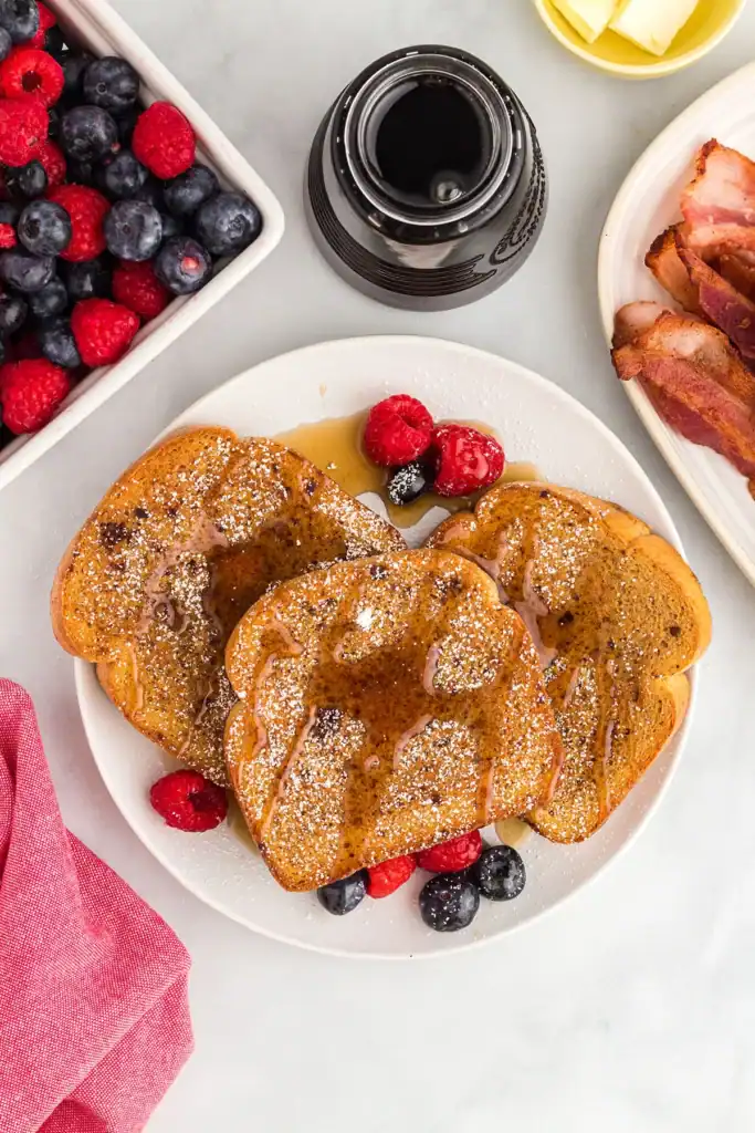 Three slices of French toast with powdered sugar, syrup, and berries on a white plate. Bowl of berries, bacon, syrup bottle, and butter are next to the plate.