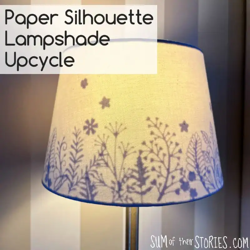 A lampshade decorated with a paper silhouette of plants and flowers, casting intricate shadow patterns when the lamp is lit.