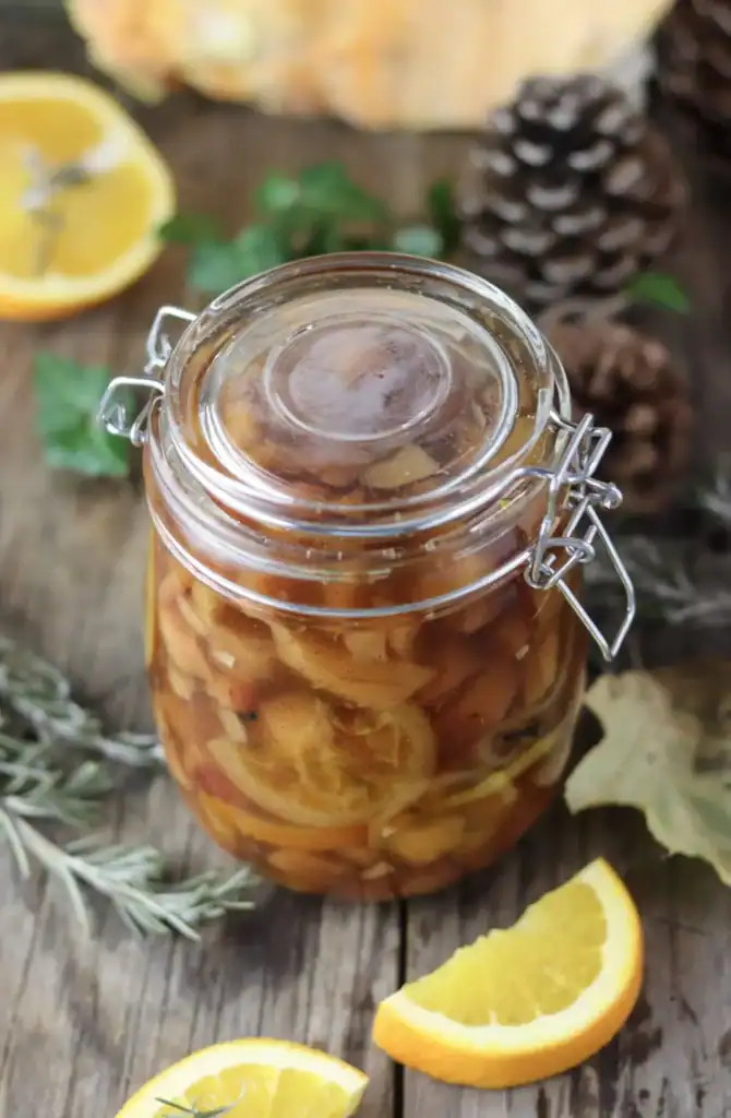 A glass jar filled with preserved fruit slices, mainly orange and apple, sitting on a wooden surface with orange wedges, rosemary, pinecones, and leaves nearby.