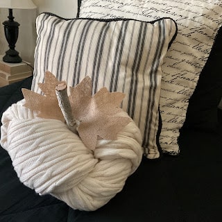 A decorative white yarn pumpkin with burlap leaves and a stick stem sits in front of two pillows, one striped and one with script text, on a dark surface.