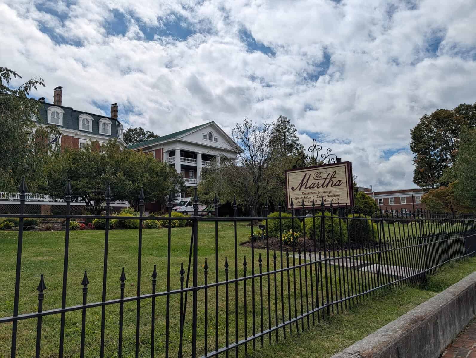 A large historic building with columns and dormer windows sits behind a black iron fence and a sign reading "The Martha." The sky is partly cloudy.