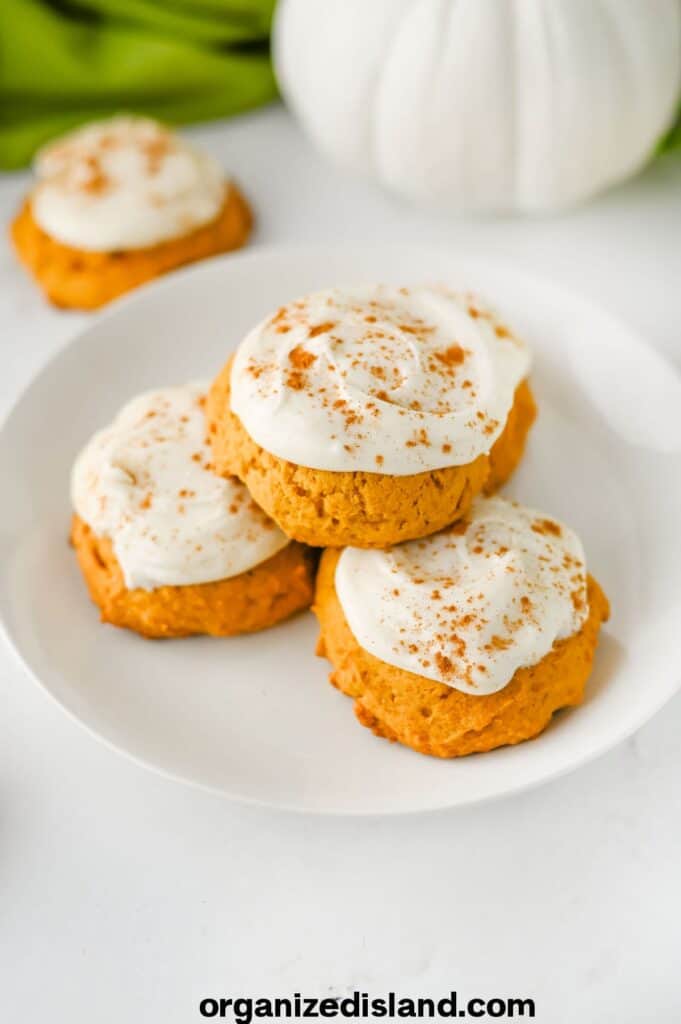 Three pumpkin cookies with white frosting and cinnamon sprinkled on top are arranged on a white plate, with two more cookies and a white pumpkin in the background.