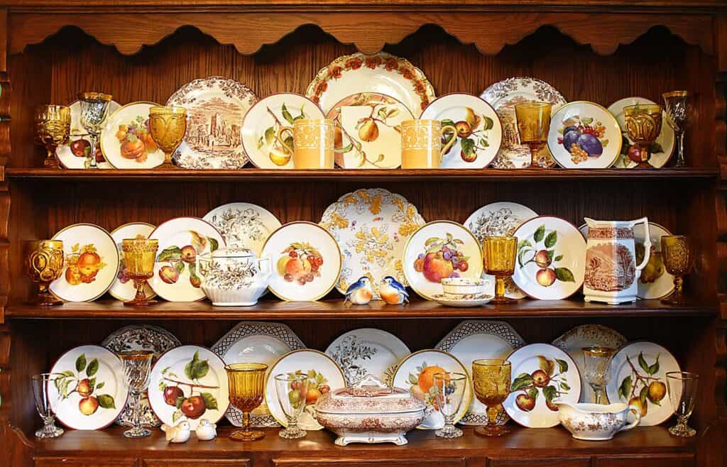 A wooden hutch displays decorative plates with fruit designs, amber glassware, ceramic figurines, and ornate serving dishes arranged on three shelves.