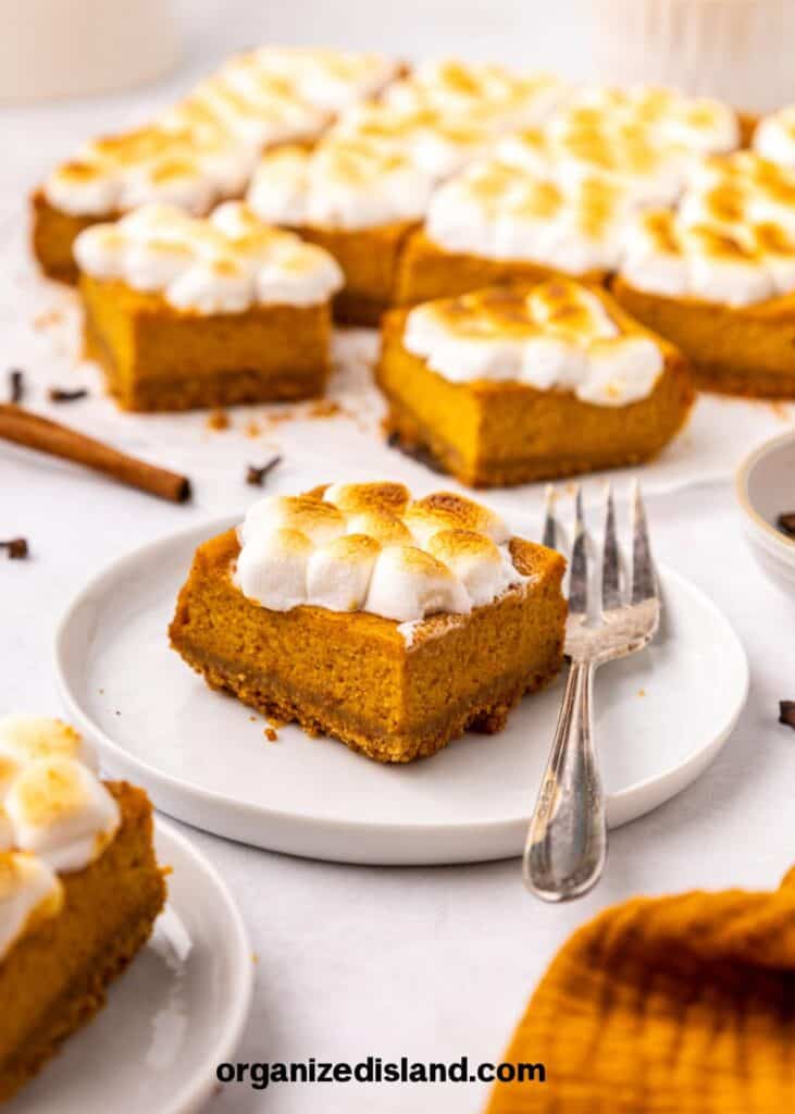 A plate with a pumpkin dessert bar topped with toasted marshmallows, a fork, and more bars in the background.