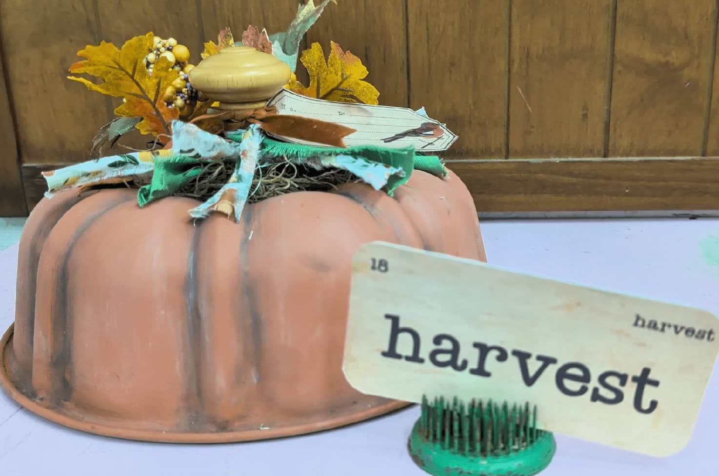 A decorative pumpkin centerpiece with artificial leaves and a "harvest" card on display in front of a wooden background.