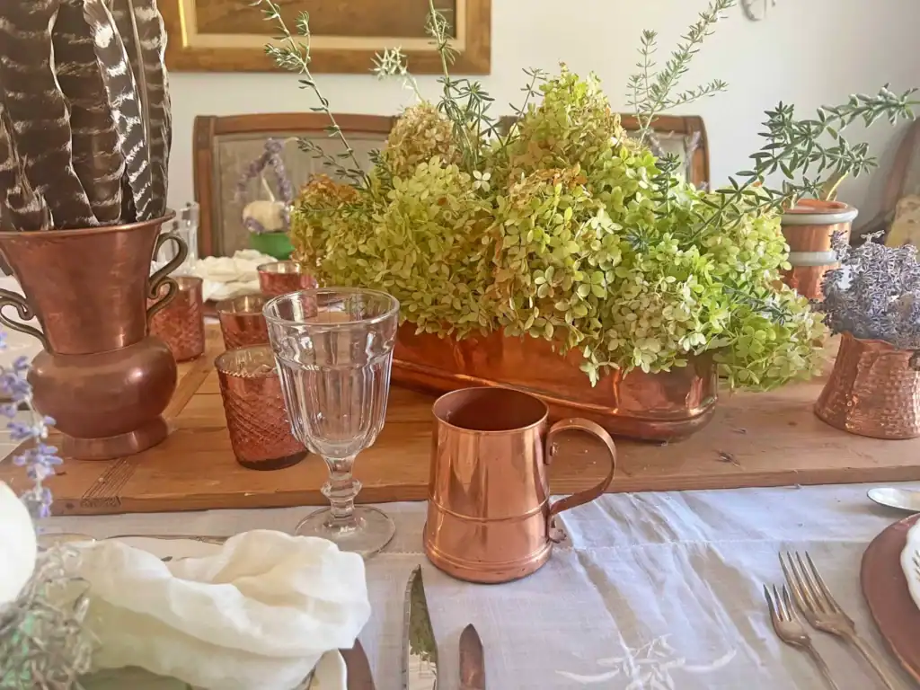 A wooden table set with copper vases, a glass goblet, a copper mug, and a centerpiece of green hydrangeas and greenery in a copper planter.