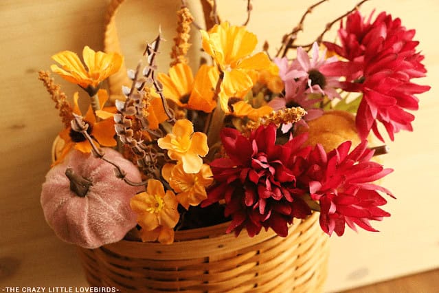 A woven basket filled with artificial autumn flowers in yellow and red, alongside a small fabric pumpkin, displayed against a light wooden background.