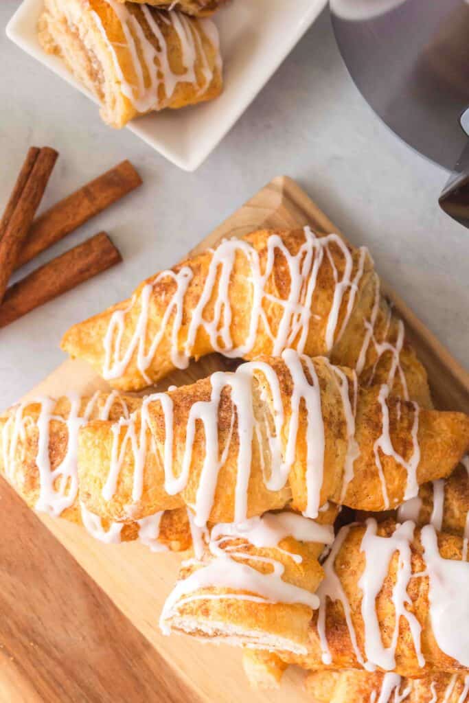 Croissants drizzled with white icing are stacked on a wooden board, with cinnamon sticks and more croissants on a plate in the background.