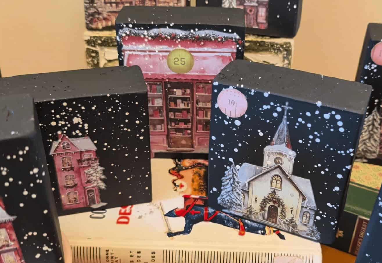 Small painted wooden blocks feature snowy village buildings, numbered windows, and festive details, arranged atop stacked books.