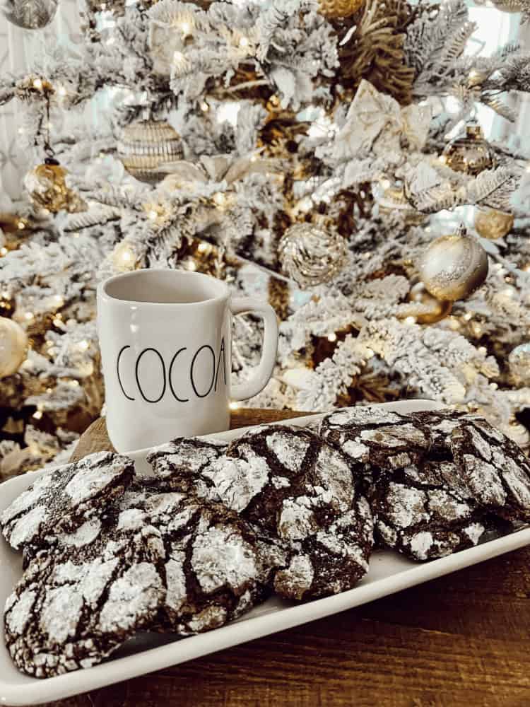 A plate of powdered chocolate crinkle cookies and a mug labeled “COCOA” sit in front of a decorated, snow-covered Christmas tree with gold ornaments.