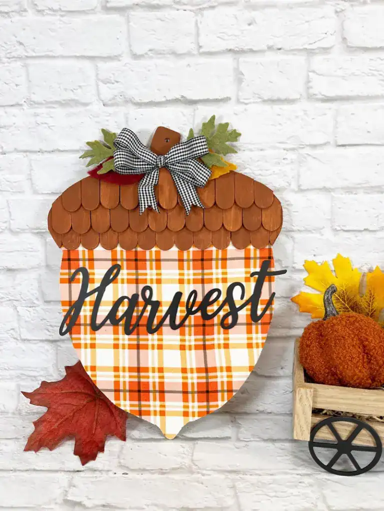 A decorative acorn-shaped sign with "Harvest" written on it, featuring a plaid pattern, bow, and leaves, displayed next to autumn leaves and a small pumpkin wagon.