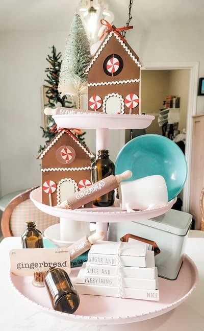 A two-tiered tray displays gingerbread house decorations, a mini Christmas tree, glass bottles, a bowl, a scoop, and books in a festive kitchen setting.