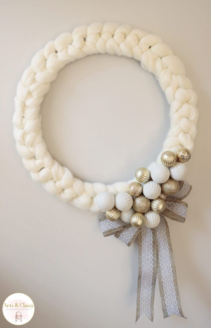 A thick, braided white wreath decorated with gold and white ornaments and a grey lace ribbon bow is displayed against a plain background.