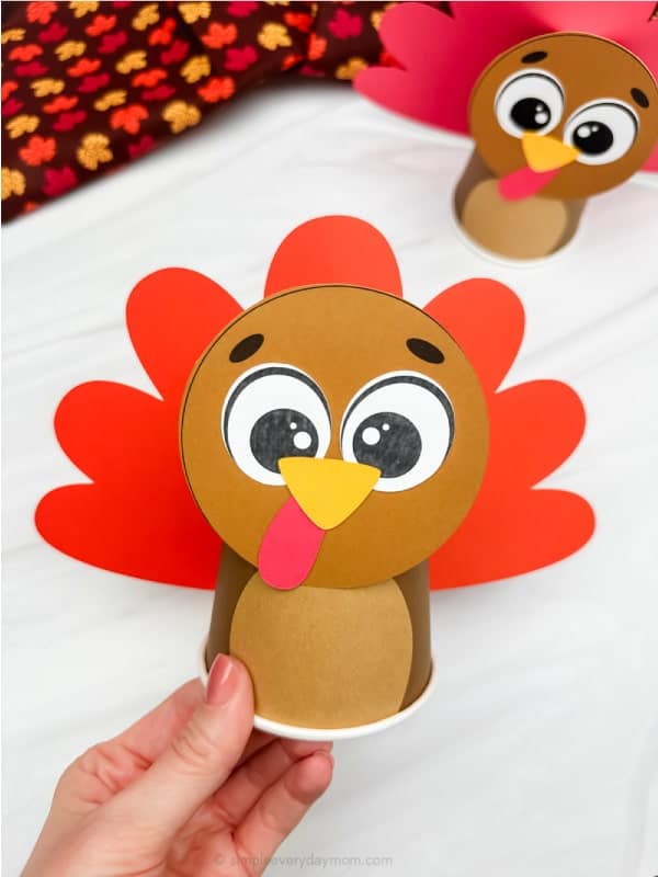 A hand holds a paper cup turkey craft with a cartoon face and red paper feathers; another similar turkey is in the background.