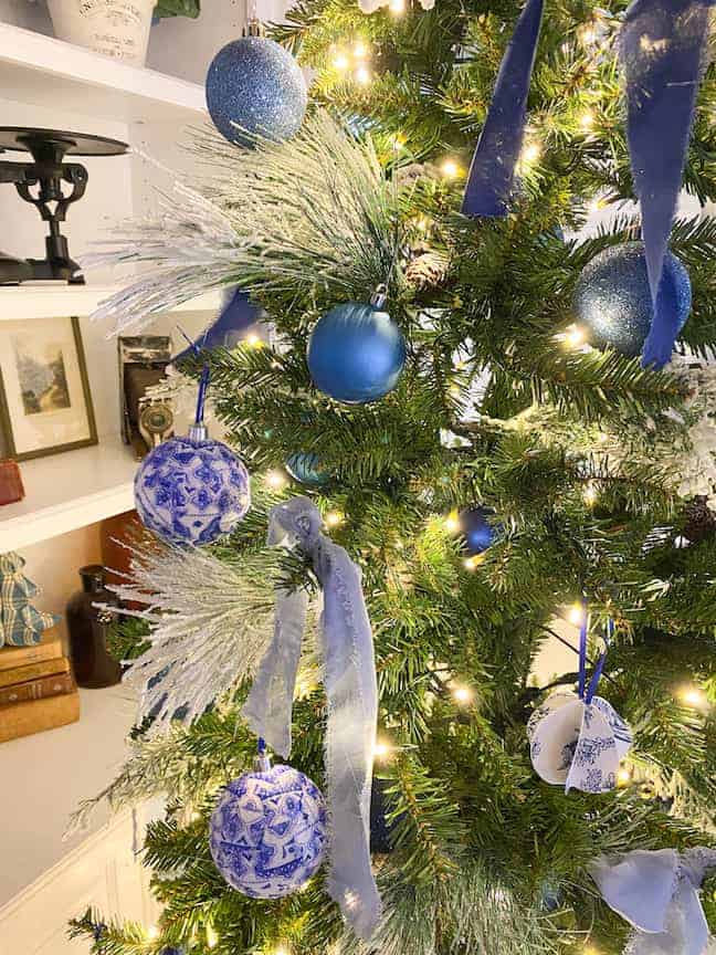 A Christmas tree decorated with blue and white ornaments, blue ribbons, and white lights stands in a room with shelves in the background.