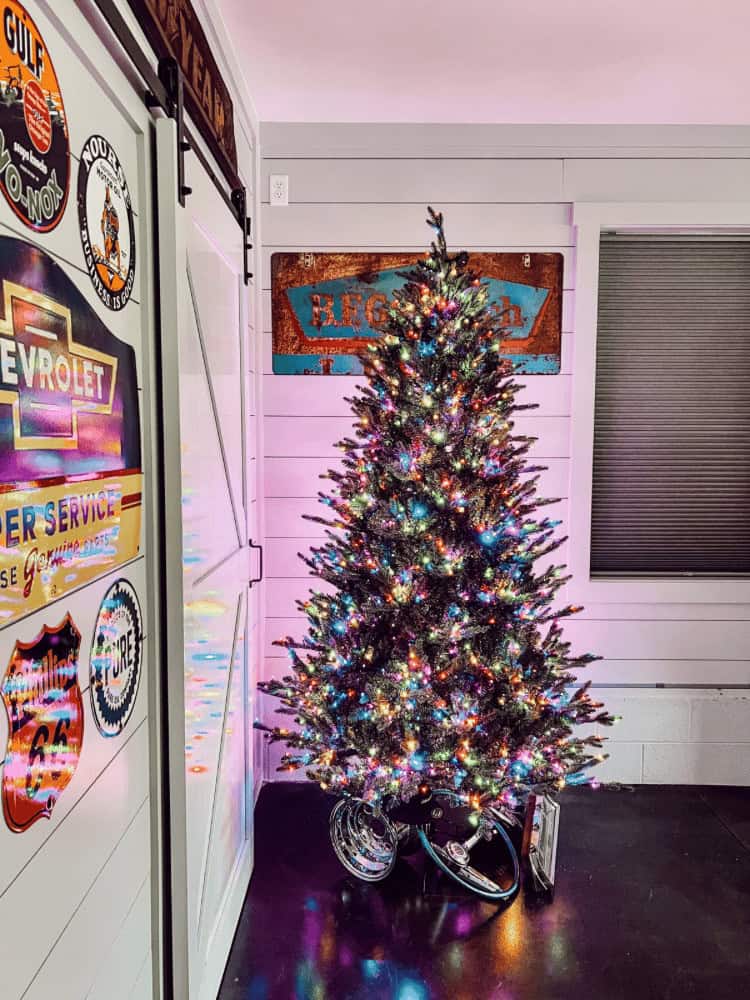 A decorated Christmas tree with multicolored lights stands in the corner of a room with vintage signs and a sliding barn door adorned with retro automotive decals.