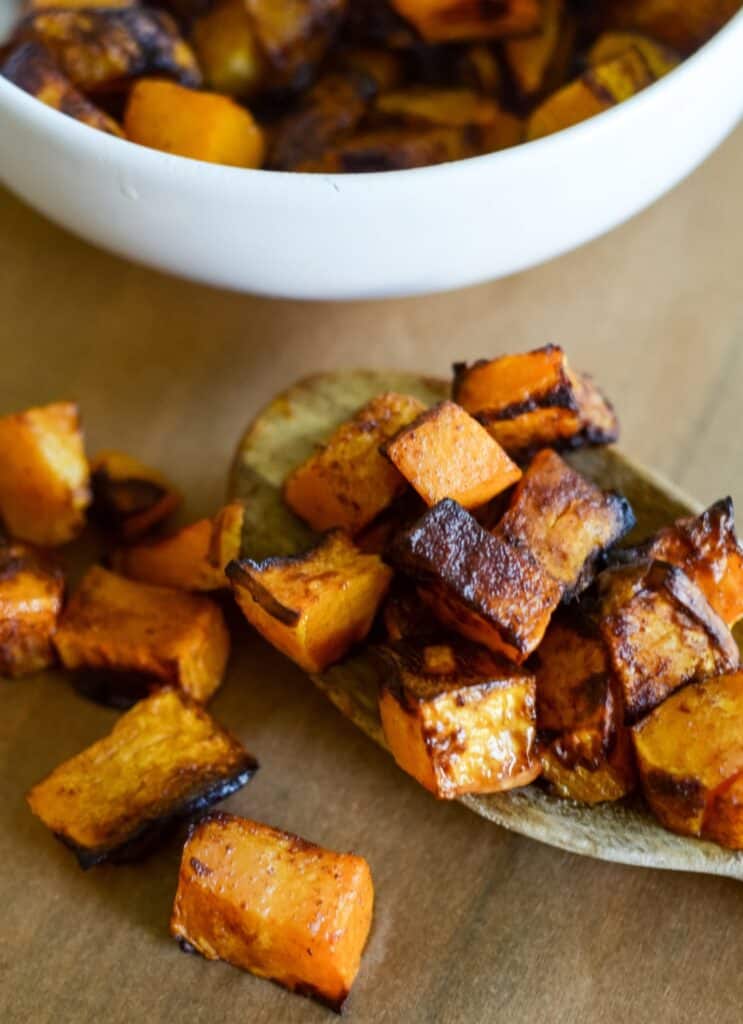 Roasted sweet potato cubes, some charred, on a wooden spoon and parchment surface, with a white bowl of more roasted potatoes in the background.