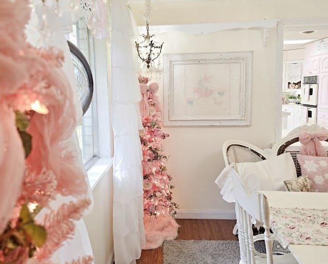 A pastel-themed room features a small pink Christmas tree, white furniture, floral accents, and soft natural light from a nearby window.